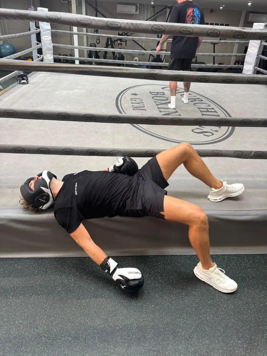 A fighter in head guard and gloves lying across the ring apron in a staged knockdown